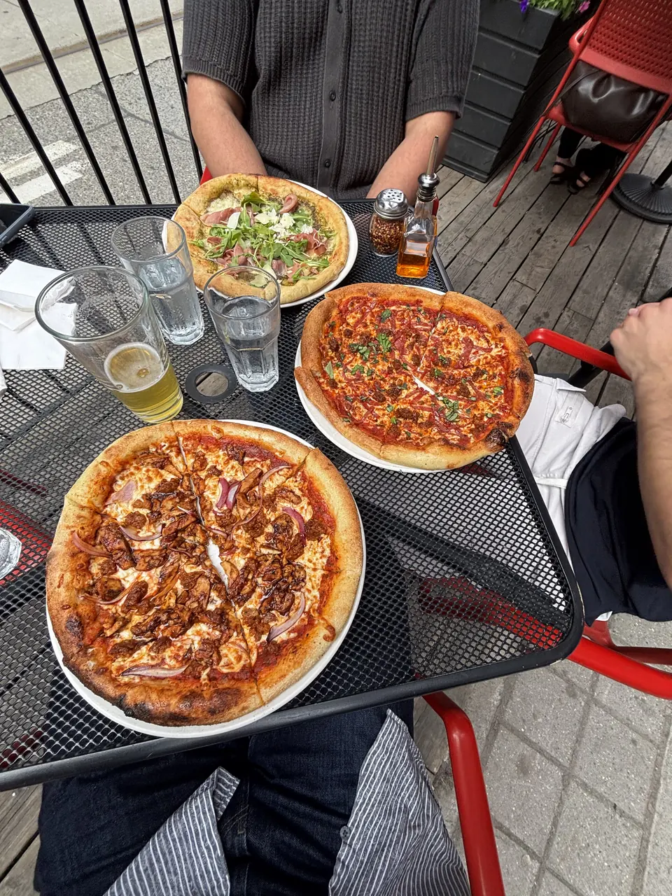 Three pizzas crowding a mesh patio table — pesto and prosciutto, one red, one with barbecue chicken and red onion